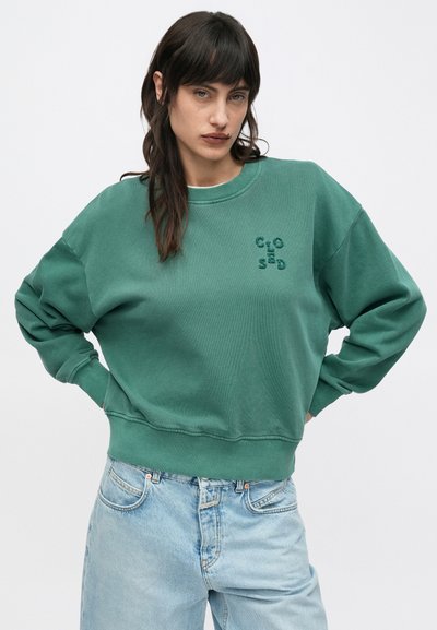 Νεαρή γυναίκα με σκούρα μαλλιά που φοράει πράσινο φούτερ με κεντημένα γράμματα και ανοιχτόχρωμο τζιν ψηλόμεσο, ποζάρει με τα χέρια στη μέση.