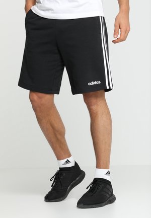 Mężczyzna ubrany w czarne krótkie spodenki Adidas z białymi paskami po bokach, białe skarpetki Adidas i czarne buty sportowe Adidas, stoi z rozluźnionymi rękami.