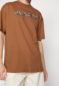 Brun bomull t-shirt med färgglad blomsterbroderi över bröstet. Korta ärmar och en klassisk rund halsringning.