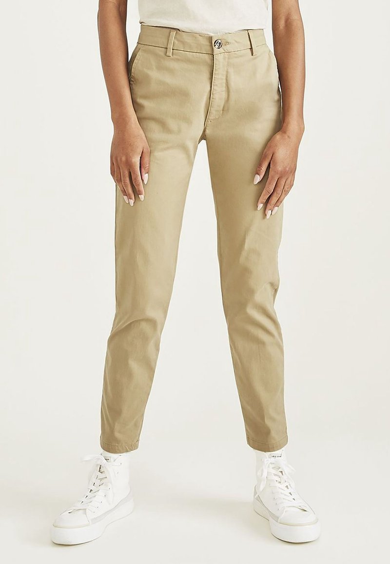 Personne portant un pantalon beige fuselé avec des poches devant et des baskets blanches montantes, debout devant un fond clair uni.