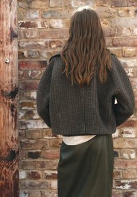 Bruin geruit cropped jasje met verlaagde schouders, gecombineerd met een lichte top en groene rok, tegen een gestructureerde stenen muur.
