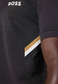 Polo noir avec un motif texturé, présentant un accent rayé or, blanc et noir près de l'épaule gauche et un logo blanc.