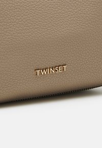 Beige textuur leren tas met een gouden "TWINSET" logo. Heeft een rits langs de rand, die het slanke ontwerp en de materiaalkwaliteit benadrukt.