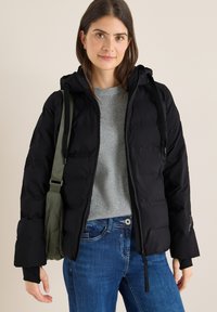 Sort quiltet pufferjakke med hætte, lange ærmer, lynlås og ribbede manchetter. Bæres over en lysegrå sweater og blå jeans.