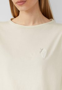 Lichtbeige katoenen t-shirt met een ronde halslijn. Heeft een witte schelpafbeelding en het woord "calm" in het grijs, minimalistisch ontwerp.