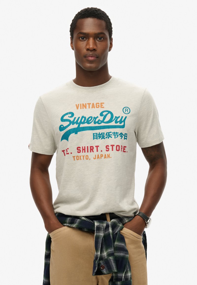Lichtgrijze katoenen t-shirt met veelkleurige tekst, met "SuperDry" in turquoise. Draag je met een geruite blouse om de taille gebonden.