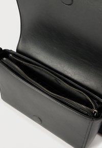 Μαύρη δερμάτινη τσάντα χιαστί με ανοιχτό καπάκι που αποκαλύπτει εσωτερικό φερμουάρ και κλείσιμο με μαγνητικό κουμπί.