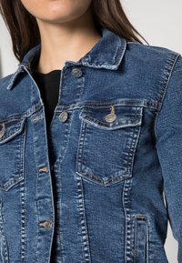Jeansjacka i medelblått med två bröstfickor, metallknappar med tryck och en klassisk krage. Har en lätt sliten textur.