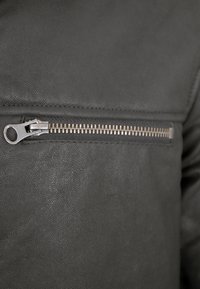 Schwarze Lederjacke mit einer diagonal verlaufenden Reißverschlusstasche und einem metallisch silbernen Reißverschluss. Die Struktur ist leicht genarbt.