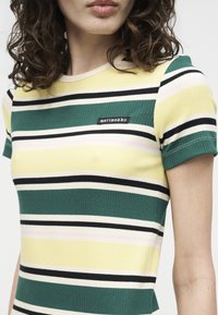 Γυναίκα που φοράει ριμπ μπλουζάκι με κοντά μανίκια, με οριζόντιες ρίγες σε πράσινο, κίτρινο, μαύρο και λευκό, και μια μικρή ετικέτα «marimekko» στο στήθος.