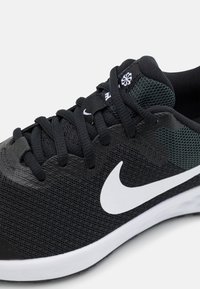Zapatilla deportiva negra con parte superior de malla, logotipo swoosh blanco en contraste, puntera negra texturizada y diseño de cordones. Acentos verdes en el talón.