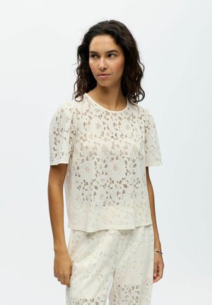 Femme aux cheveux bouclés foncés portant un haut court en dentelle florale blanche et un pantalon assorti, debout devant un fond uni.