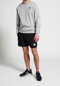 Uomo in felpa a maniche lunghe grigio chiaro, pantaloni corti neri e scarpe da ginnastica nere e blu che si trova contro uno sfondo bianco.