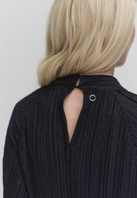 Mørkeblå top med et tekstureret materiale, forsynet med en åbning i nakken, der lukkes med en cirkulær metaldetalje.