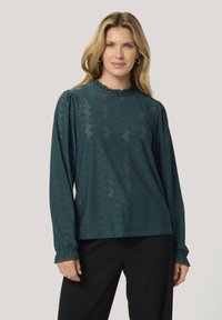 Tropfengrünes Langarm-Blusenshirt mit einem gerafften Ausschnitt, das mit kunstvollen Ausstanzungen und geknöpften Manschetten versehen ist. Kombiniert mit dunklen Hosen.