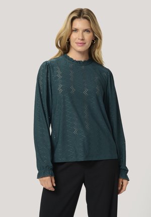 Tropfengrünes Langarm-Blusenshirt mit einem gerafften Ausschnitt, das mit kunstvollen Ausstanzungen und geknöpften Manschetten versehen ist. Kombiniert mit dunklen Hosen.