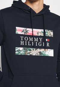 Granatowa bluza z kapturem z motywem palm i zachodzącego słońca, napisem „TOMMY HILFIGER” oraz logo marki w postaci czerwono-biało-niebieskiej flagi na piersi.