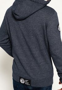 Ciemnoszara bluza z kapturem, z prążkowanymi mankietami i dołem. Na lewym rękawie znajduje się okrągłe logo, a z tyłu etykieta.