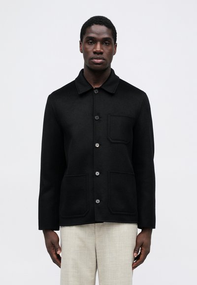 Veste en laine noire avec col, cinq boutons et deux poches avant, présentant une texture douce et une silhouette cintrée.