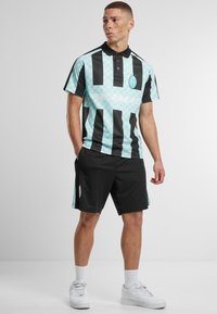 Polo shirt in lichtblauw met zwarte strepen; voorzien van een kraag, korte mouwen en een cirkelvormig logo. Gecombineerd met zwarte shorts en witte sneakers.