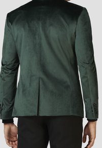 Zielona aksamitna marynarka o gładkiej teksturze, z pojedynczym rozcięciem z tyłu i guzikami na rękawach, połączona z czarnymi spodniami.
