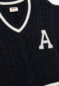 Granatowy sweter robiony na drutach z wzorem w warkocze, z dekoltem w kształcie V i kremowym ściągaczem. Ma na przodzie kontrastującą białą literę "A".