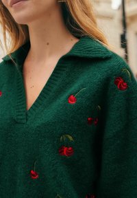 Green knitted sweater with a V-neck, featuring red cherry embroidery and a textured finish. The collar is turned down, accentuating the design.
