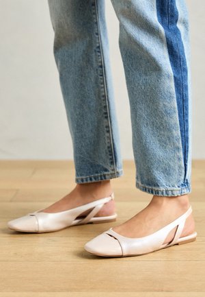 Stopy w różowych jasnych balerinach typu slingback z szpiczastym noskiem i wycięciami, w połączeniu z podwiniętymi dwukolorowymi jeansami na drewnianej podłodze.