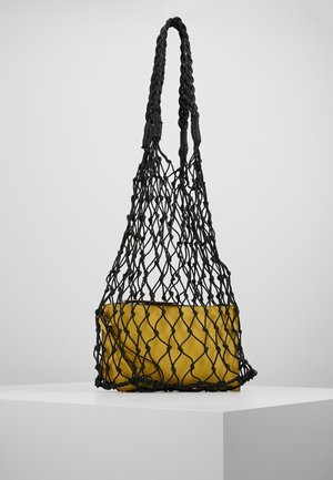 Czarna pleciona siatkowa torba zawierająca prostokątną, musztardowo-żółtą saszetkę, ustawiona na białym postumencie na tle jednolitego szarego tła.