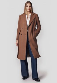 Podwójnie zapinany brązowy płaszcz wełniany z klapami, bocznymi kieszeniami i czarnymi guzikami, noszony na białej koszuli i niebieskich dżinsach.