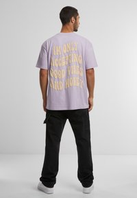 Lyse lilla oversized T-shirt med stor gul tekst på ryggen. Bæres med sorte cargo-bukser og hvide sko. Neutral baggrund.