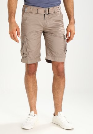Mand iført beige cargo shorts til knæet med sidelommer, beige bælte, hvide sneakers og blå t-shirt, står mod en hvid baggrund.