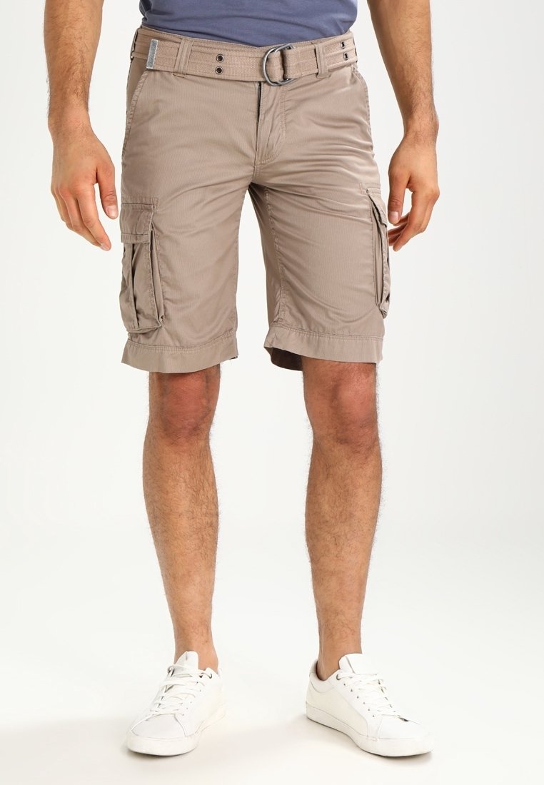 Homme portant un short cargo beige jusqu'aux genoux avec des poches latérales, ceinture beige, baskets blanches et t-shirt bleu, debout devant un fond blanc.
