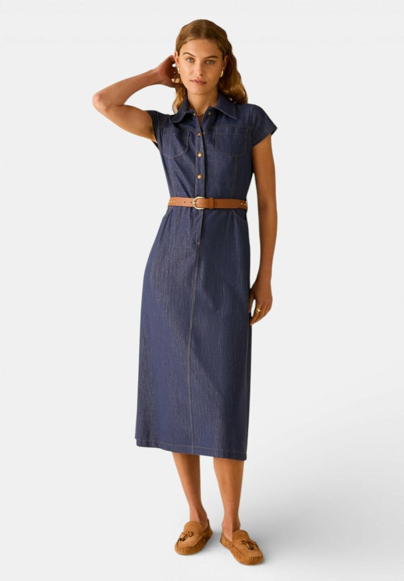 Giovane donna che indossa un abito midi senza maniche in denim scuro con bottoni frontali e una cintura marrone, abbinato a mocassini color cuoio, che posa con una mano dietro la testa.