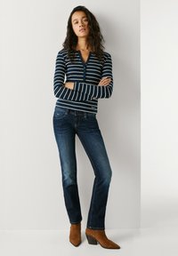 Donna con lunghi capelli ricci in piedi con le braccia incrociate, indossa una camicia a righe a maniche lunghe, jeans blu scuro e stivaletti marroni.
