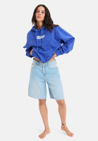 Sudadera de algodón azul con bolsillo frontal y gráfico blanco; combinada con pantalones cortos de mezclilla azul claros. El modelo está descalzo, con los brazos a los lados.
