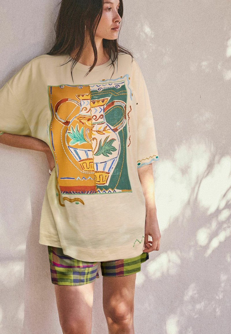 Femme portant un T-shirt beige oversize avec une illustration colorée de vase et un short vert à carreaux, debout contre un mur clair texturé avec des ombres.