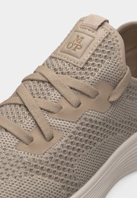 Μπεζ πλεκτό αθλητικό παπούτσι με υφαντή υφή, κορδόνια που ταιριάζουν και λογότυπο στην γλώσσα, με λευκή σόλα με αυλάκια.