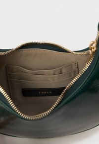 Zaļas ādas rokassomu iekšpuse ar bežīgu oderi, kurā ir karšu slots un zelta rāvējslēdzis. Zīmola uzlīme iekšpusē norāda "Furla Since 1927."