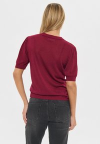 Burgundska pletena pulover s kratkimi napihnjenimi rokavi in ribastim robom, v kombinaciji s sivimi jeans hlačami z zadnjimi žepi. Gladka tekstura in oprijet kroj.