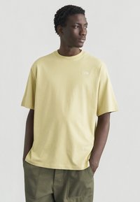 Ανοιχτό κίτρινο βαμβακερό μπλουζάκι με στρογγυλή λαιμόκοψη και κοντά μανίκια· διαθέτει μικρό λευκό λογότυπο στο αριστερό πάνω μέρος του στήθους.