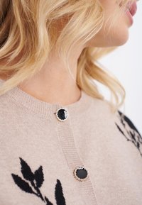 Beige cardigan med svart blommönsterbroderi, med runda svarta knappar med silverdetaljer. Mjuk textur och klassisk rund halsringning.