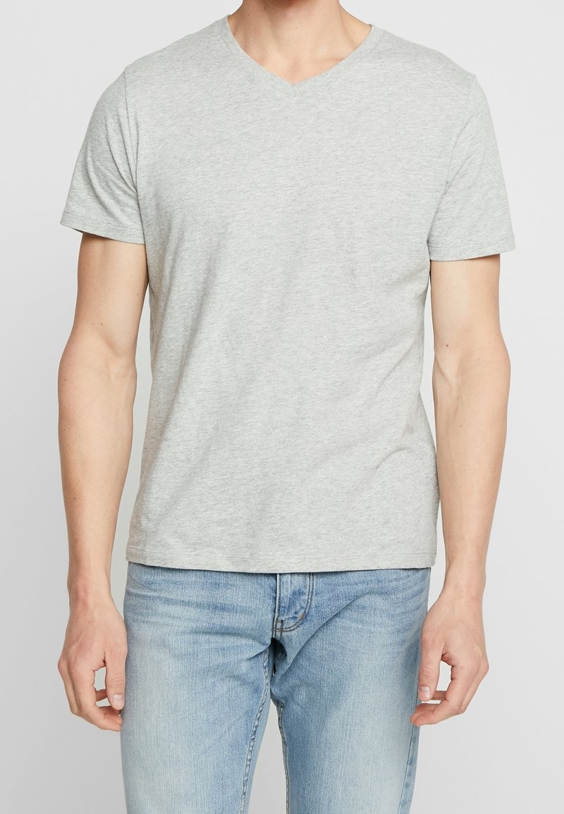 Homme portant un T-shirt V-cou gris clair et un jean bleu clair, se tenant avec les bras relaxés sur les côtés devant un fond uni.