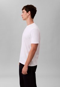 Biała koszulka z krótkim rękawem i okrągłym dekoltem, wykonana z gładkiej tkaniny, noszona przez męskiego modela na szarym tle; zestawiona z ciemnymi dżinsami.