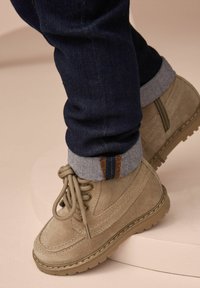 Beige suède enkellaarzen met ronde neus, vetersluiting en rubberen zolen. Gecombineerd met donker jeans en omgeslagen manchet dat denim en oranje stiksels laat zien.