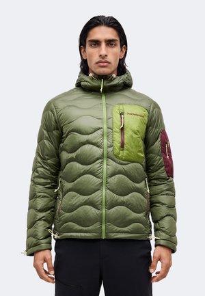 Man draagt een groene doorgestikte jas met capuchon, lichtgroene borstzak, kastanjebruine mouwzak en zwarte broek, staand tegen een effen achtergrond.
