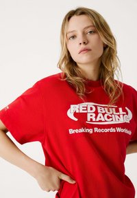 Punainen lyhythihainen t-paita, jossa on valkoisella tekstillä "RED BULL RACING" ja "Breaking Records Worldwide". Valmistettu pehmeästä puuvillakankaasta.