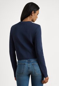 Tumši zils īss džemperis ar garām piedurknēm, teksturētu adījuma audumu, apaļu kakla izgriezumu, kombinēts ar tumši izgāztiem pieguļošiem džinsiem ar aizmugures kabatām.