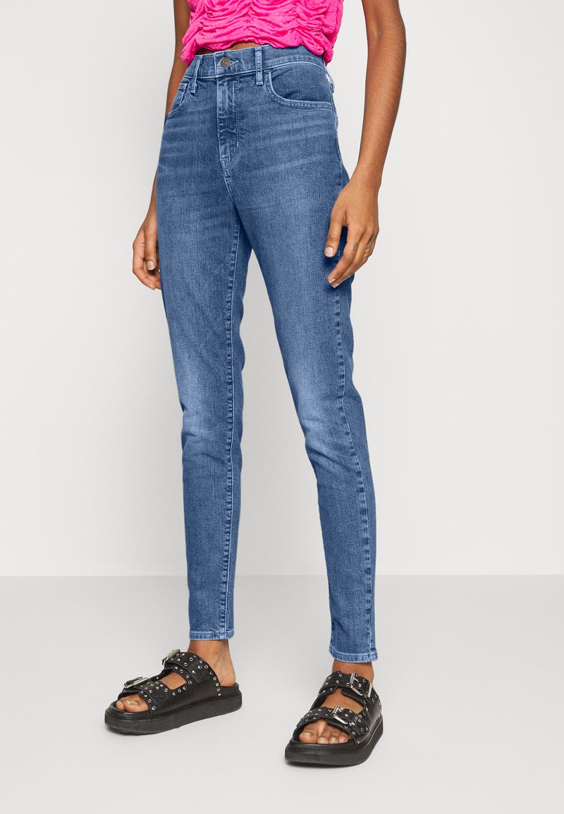 Skinny-fit blue denim jeans with a faded wash, high waist, and classic five-pocket design. Paired with black sandals featuring decorative studs.