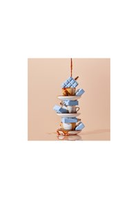 Stabel av hvite keramiske kopper med blå detaljer, toppet med blå geometrisk emballasje, kaffe som spruter over, på en reflekterende overflate.